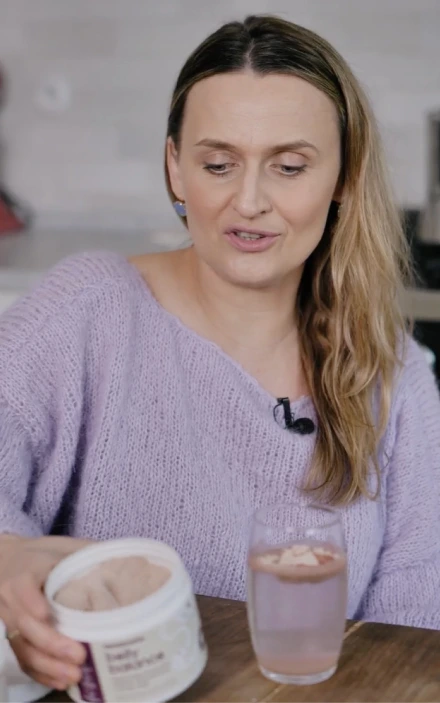 Kobieta z blond włosami ubrana w jasnofioletowy sweter z dzianiny, siedząca przy drewnianym stole z pojemnikiem suplementu diety BellyBalance i szklanką jasnoróżowego napoju.