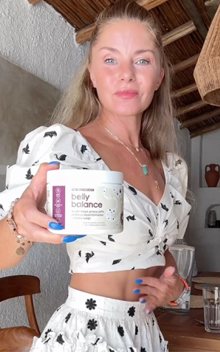 Kobieta z blond włosami ubrana w biały krótki top i spódnicę z czarnym wzorem kwiatowym, trzymająca pojemnik suplementu diety BellyBalance w rustykalnym wnętrzu z odsłoniętymi drewnianymi belkami stropowymi i białymi ścianami.