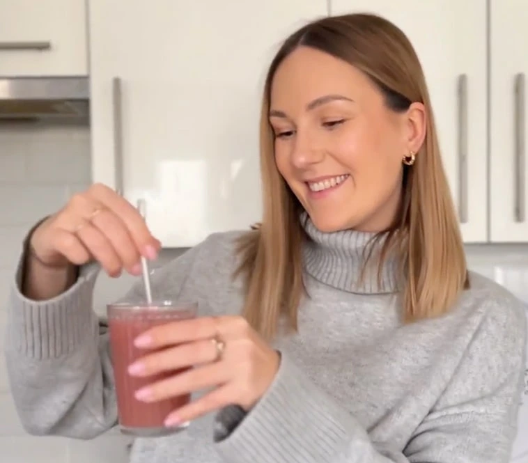 Uśmiechnięta kobieta z blond włosami ubrana w szary sweter z golfem, mieszająca łyżeczką suplement diety BellyBalance w białej kuchni.